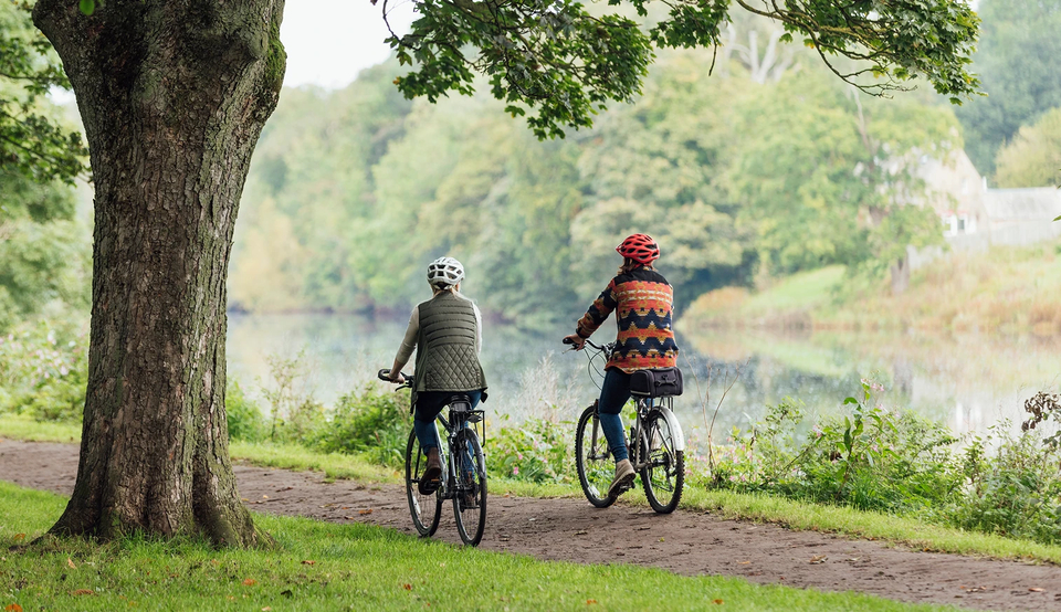 Two women, seen from behind, riding bicycles along the bank of a river.